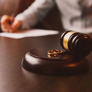 A gavel and wedding rings on a desk with a person writing in the background - Daniel Ogbeide Law.