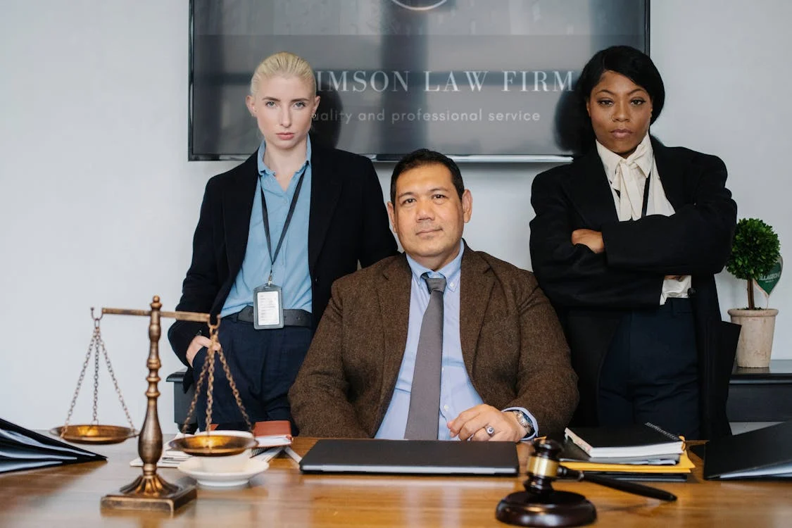 Three confident legal professionals at a desk with scales of justice, documents, and a gavel in front of them.
