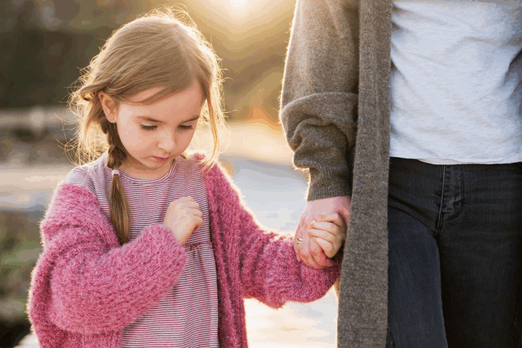 a child looking sad at the ground holding a parents hand
