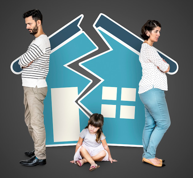 A child sitting on the floor with her parents standing on the sides of a broken house made up of cardboard