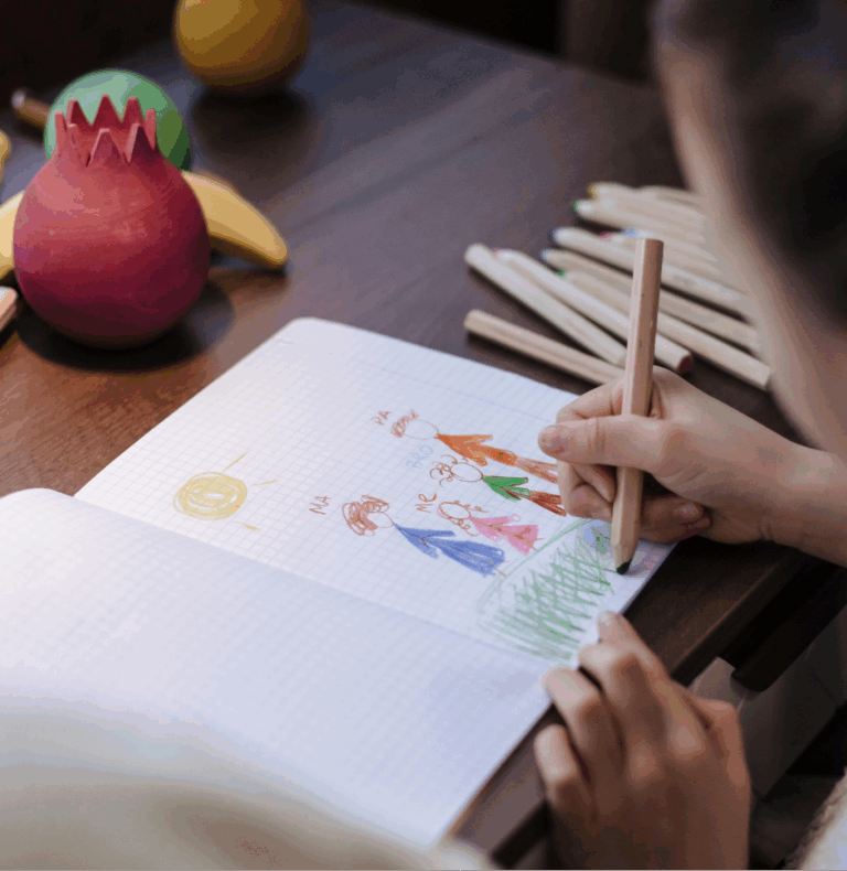 The image shows a person drawing with colored pencils in a notebook, with colorful fruit-like objects and crayons scattered on the table.