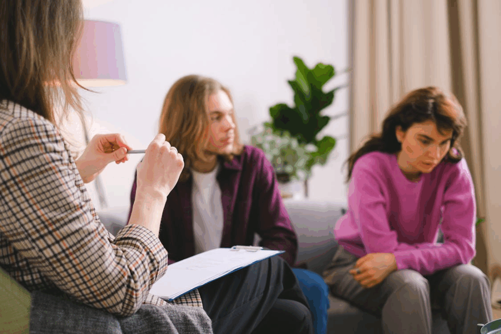 The image shows a couple sitting together, looking concerned during a therapy or counseling session, with a professional holding a notepad.