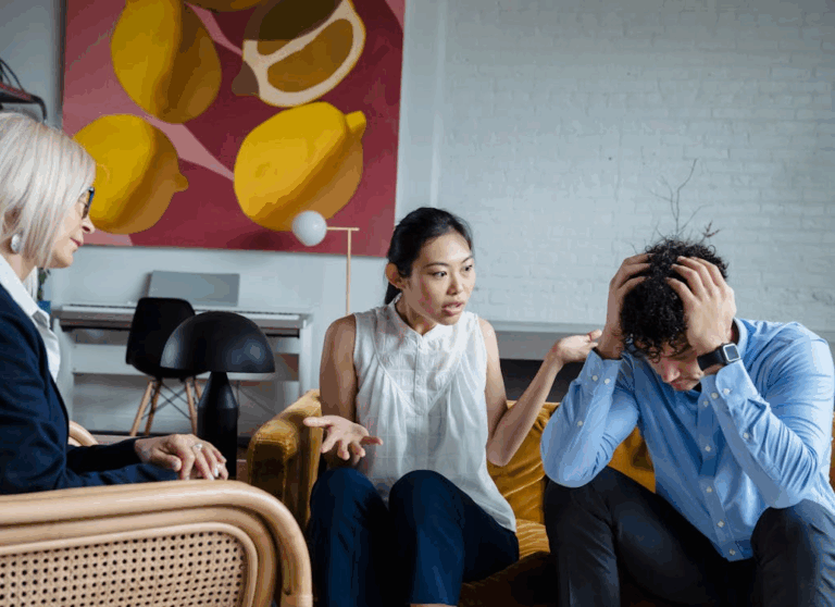The image shows a counseling or therapy session with two individuals engaged in a conversation while a professional looks on, offering support.