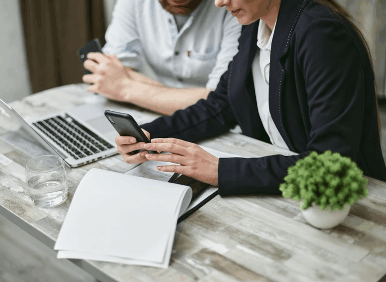 The image shows two people sitting at a table, one holding a smartphone and the other using a laptop, with documents and a small plant on the table.