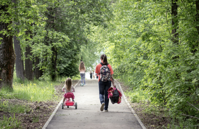 This photo shows a parent walking with a child along a park pathway while carrying the child’s backpack.