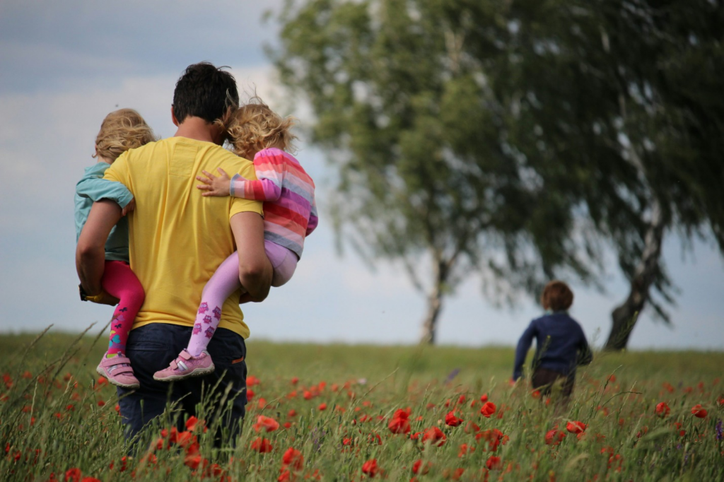 A father holding two children as one child runs in the field
