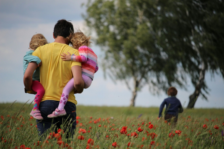 A father holding two children as one child runs in the field