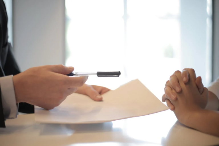 Businessman handing a contract to a woman for signing at a desk.