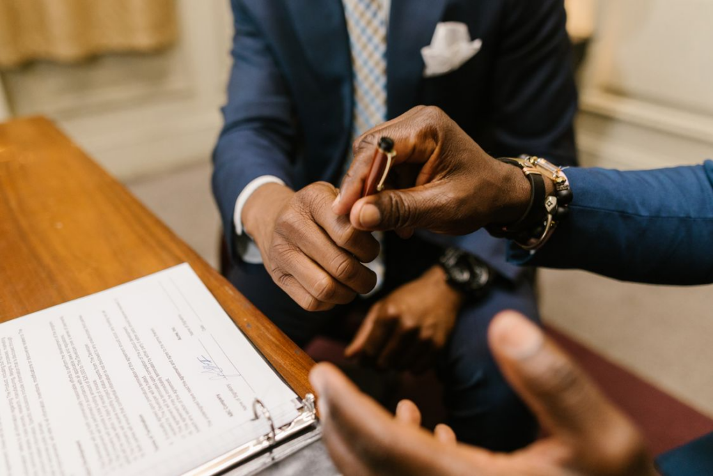 Two people at a table, one handing a pen to the other to sign documents