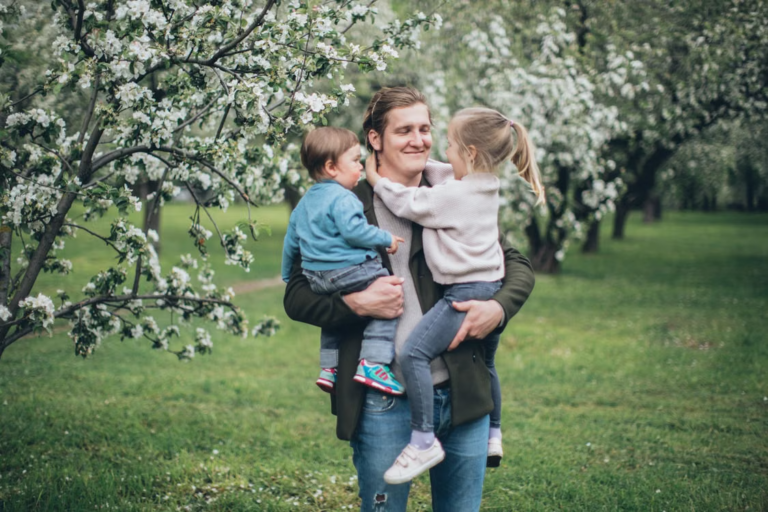 This image shows a father holding his two children outdoors near blossoming trees.
