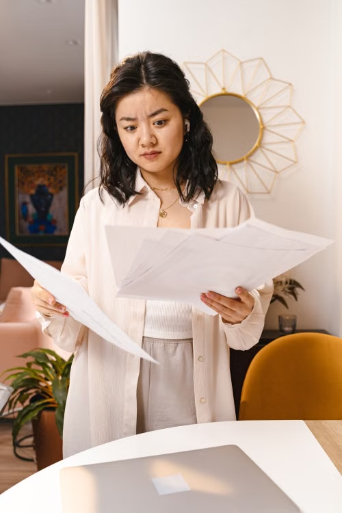 This image shows a woman looking confused while reviewing documents at home.