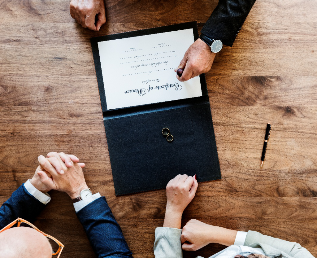 Overhead view of hands near an open certificate document with two rings and a pen on a wooden table